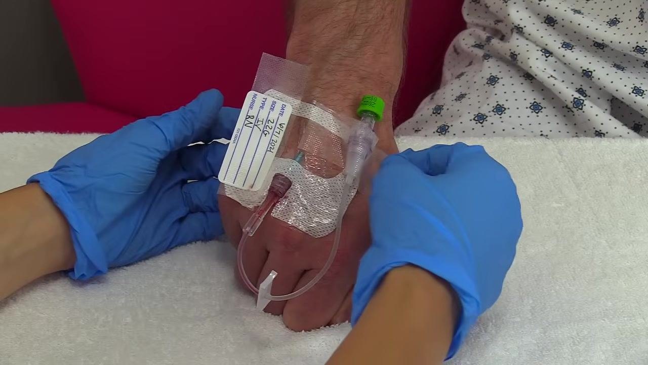 Gloved hands continue to remove tape from the IV site, stabilizing the IV