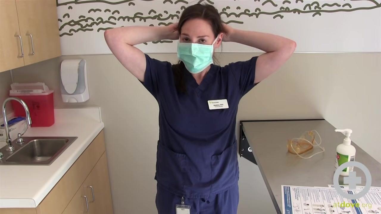 Person in scrubs pulling the gown down by the cuffs, turning it inside out, in a clinical room with a sink, sanitizer, PPE removal instructions, and goggles on the counter.