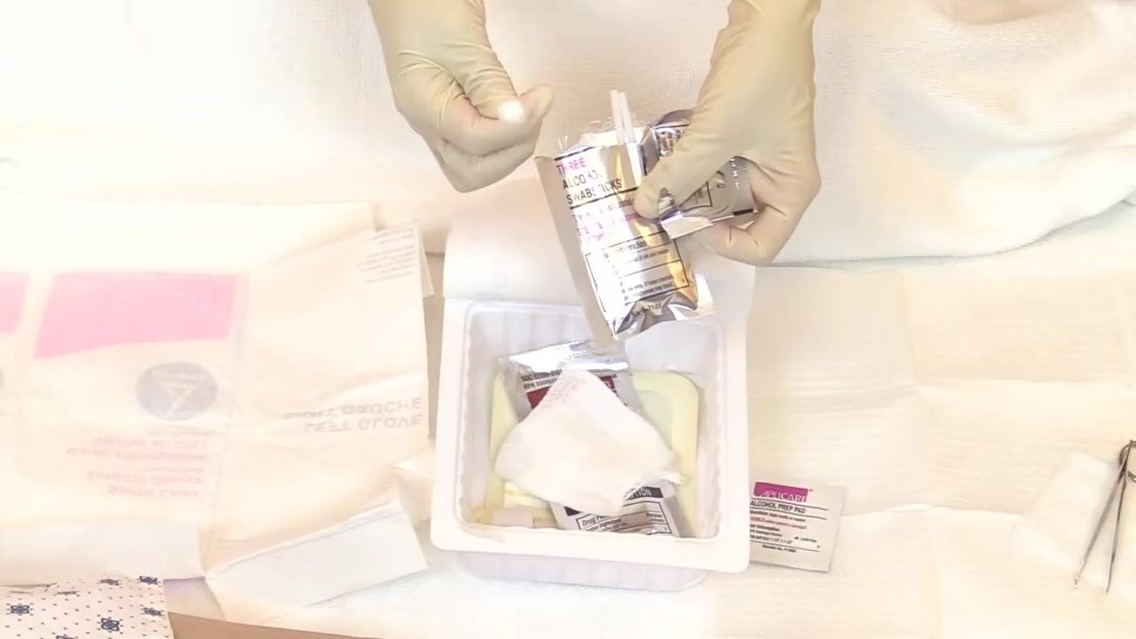 Both hands, now gloved, point to and handle antiseptic swab packages arranged in a tray with other wound care supplies.
