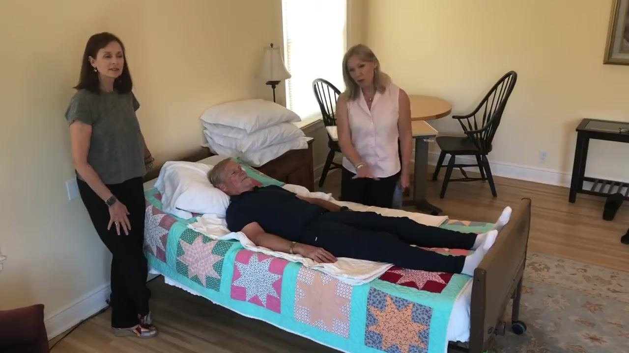 A patient lies in bed with two caregivers standing nearby. The patient's feet are pressed against the footboard, and a draw sheet is visible under the patient. The room is tidy with minimal clutter, and extra pillows are stacked on a bedside table.