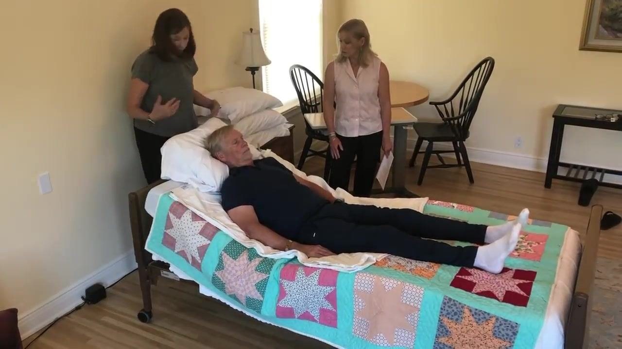 Two caregivers stand near the head and side of the bed with the patient lying comfortably. The room is spacious, with a table, chairs, and a lamp visible, and the bed is pulled out from the wall demonstrating the 18-inch gap.