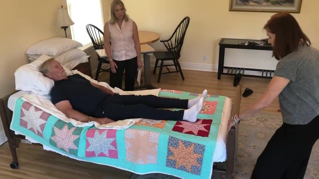 One caregiver moves the bed back toward the wall while the other observes; the patient remains in bed and the gap between the bed and the wall is visible.