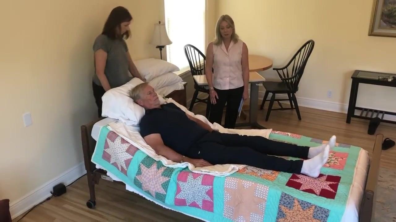 Both caregivers stand by the bed with the patient in a comfortable, elevated position. The room remains tidy and organized, with space around the bed for movement.