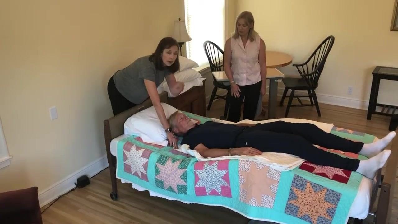 The caregiver speaks to the patient, maintaining eye contact and preparing to move them up in bed, while a second caregiver observes.