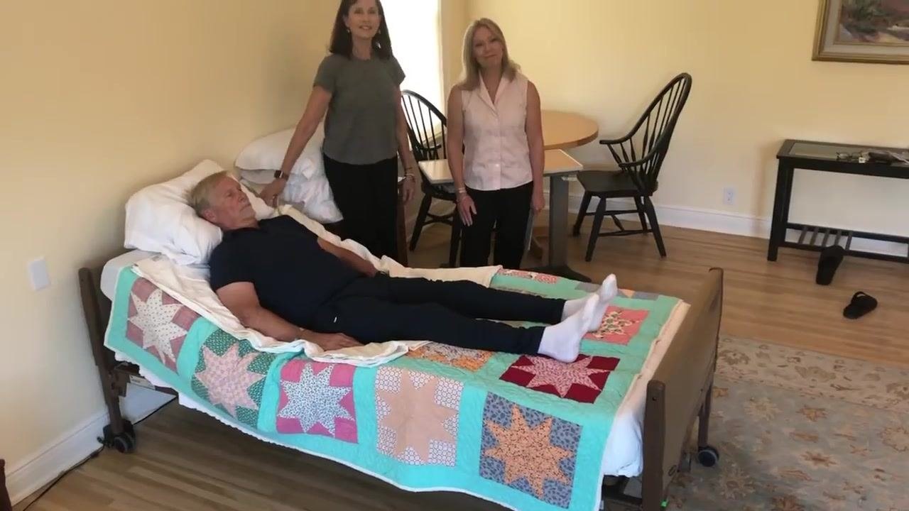 Both caregivers stand at the head and side of the bed, smiling, with the patient comfortably positioned. The bed is back in place and the room is organized and ready for ongoing care.