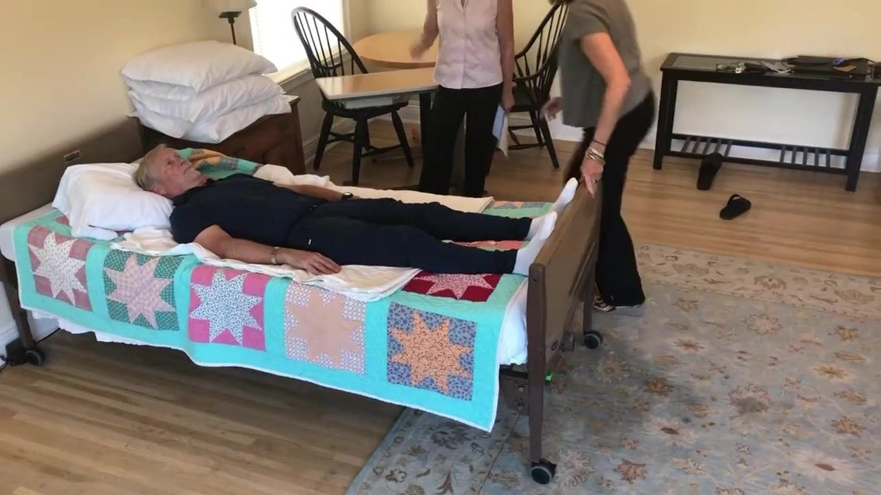A caregiver stands at the foot of the bed preparing to unlock the bed wheels, while another caregiver stands at the head of the bed. The patient remains lying flat, and the room is organized with chairs and a table in the background.