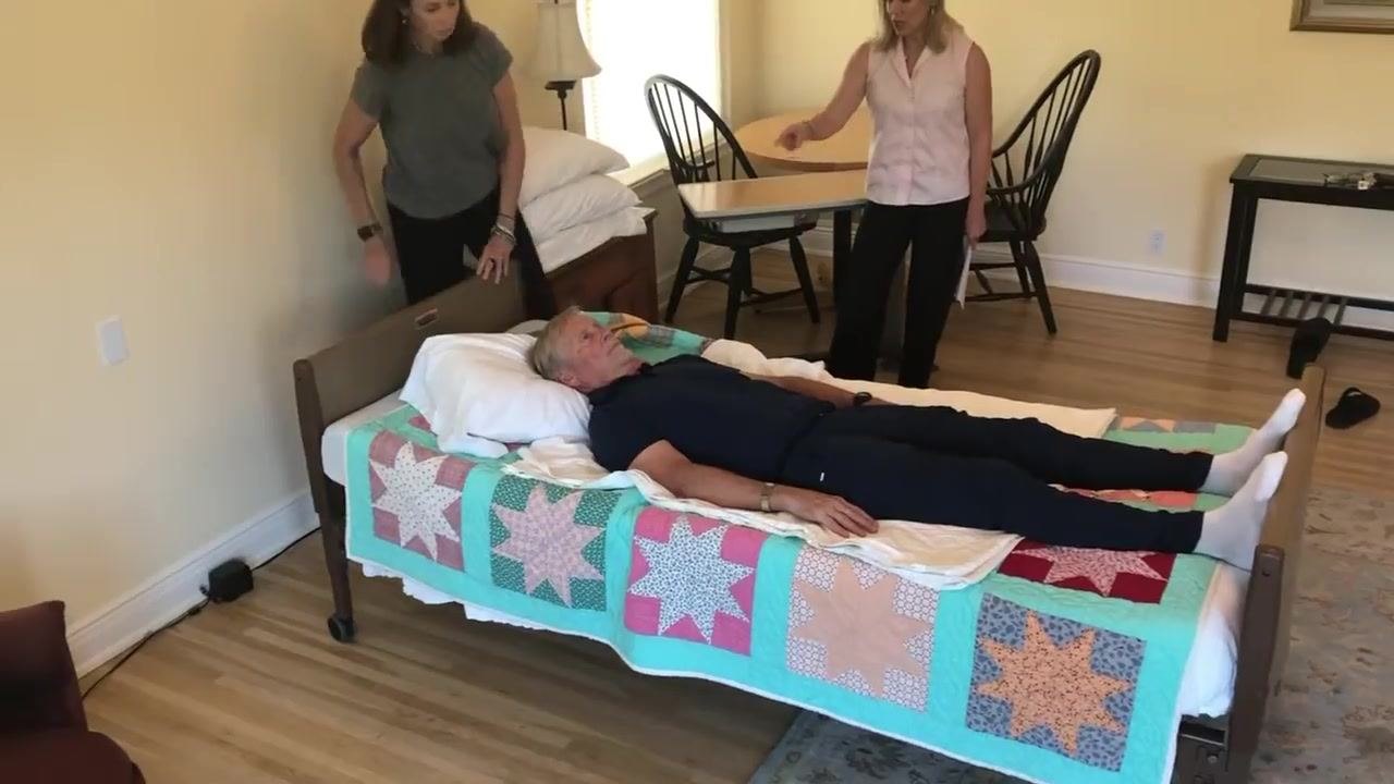 A caregiver uses the bed control to raise the foot of the bed while the patient remains lying flat. The head of the bed is not elevated, and the environment is clear and organized.