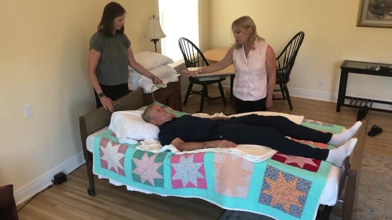 A caregiver at the head of the bed adjusts the pillow under the patient's head while a second caregiver observes nearby. The patient lies flat under a colorful quilt in a tidy room.