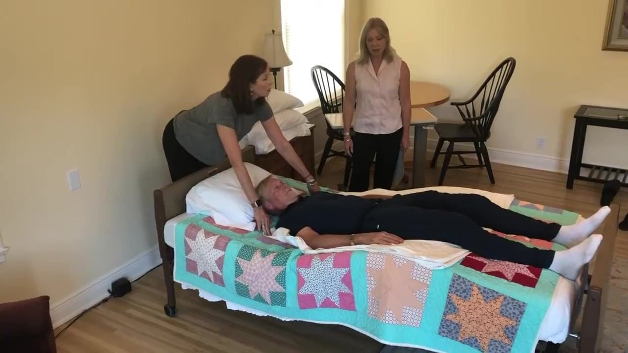 The caregiver at the head of the bed leans forward, grasping the draw sheet at the patient's shoulders in preparation for the move, while a second caregiver watches.