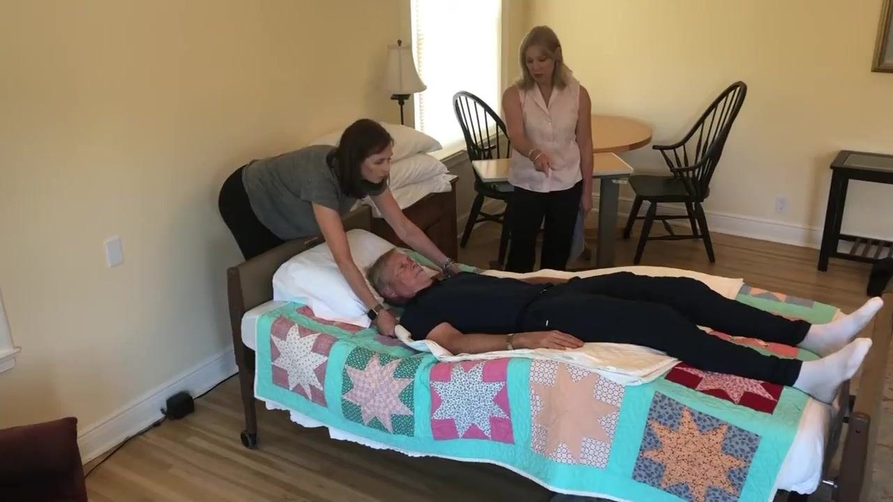 The caregiver pulls the draw sheet to move the patient up in bed while supporting the patient's head; a second caregiver observes.