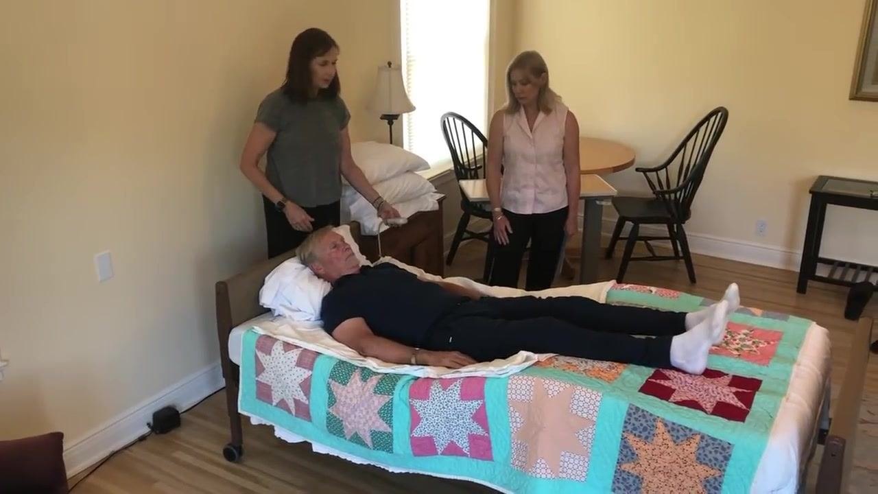 Both caregivers stand by the bed, one holding the bed control, with the patient now in a higher and more comfortable position and the bed slightly elevated at the knees.