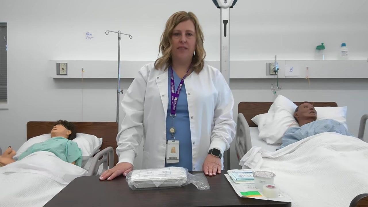A healthcare professional stands behind a table with a sealed dressing tray, tape, gauze, duoderm dressing, and cleaning solution in a clinical skills lab with two patient mannequins in hospital beds.