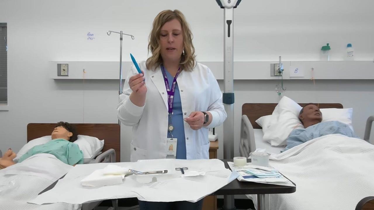 A healthcare professional holds blue forceps above the tray, showing that the handle where it was touched stays on the non-sterile border while the sterile tip remains in the sterile field. The tray contains forceps, scissors, and gauze, with the drape squared off.