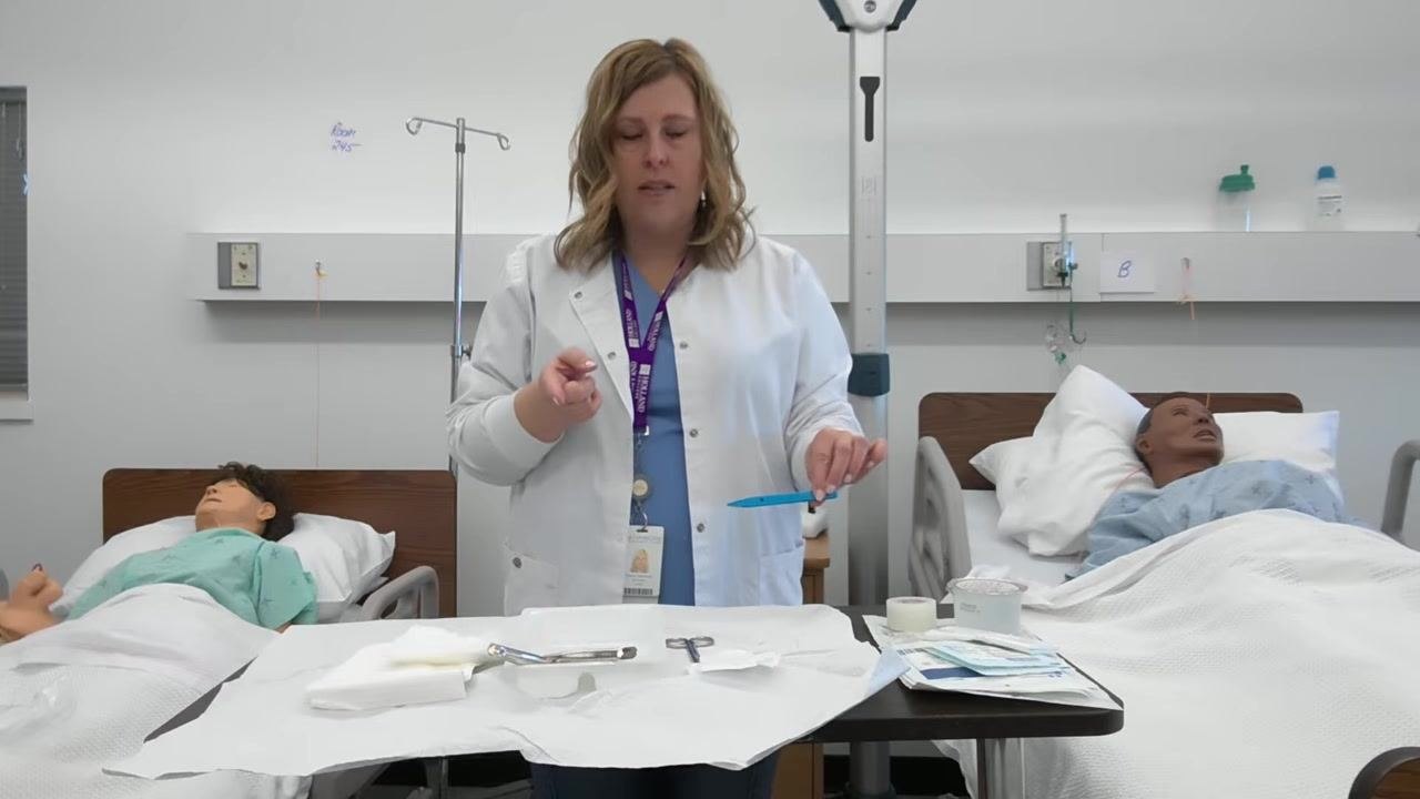 A healthcare professional holds blue forceps above a squared-off sterile tray containing forceps, scissors, gauze, and tape. Two patient mannequins are visible in the background.