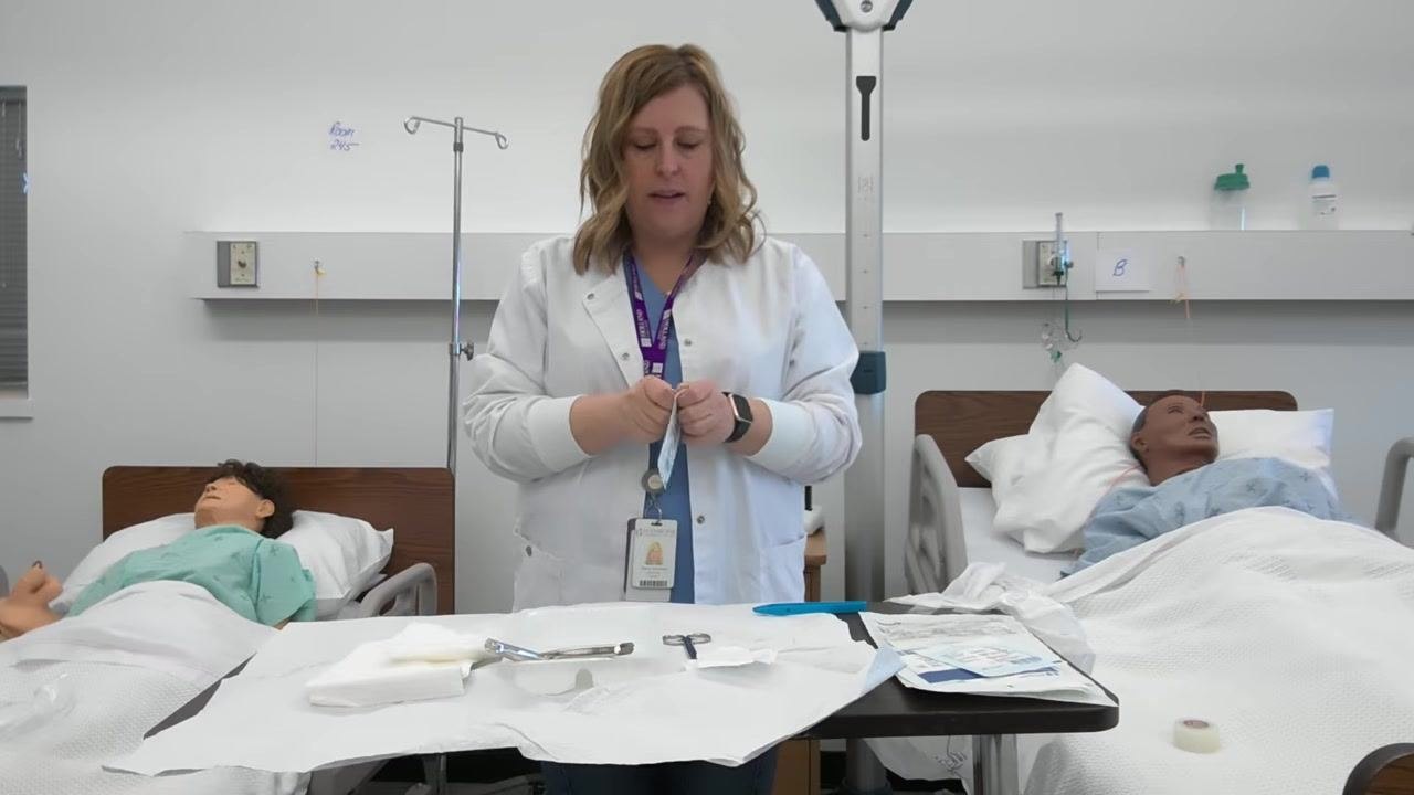 A healthcare professional peels back gauze packaging over a sterile field, with medical mannequins visible in the background and sterile instruments laid out on the tray.