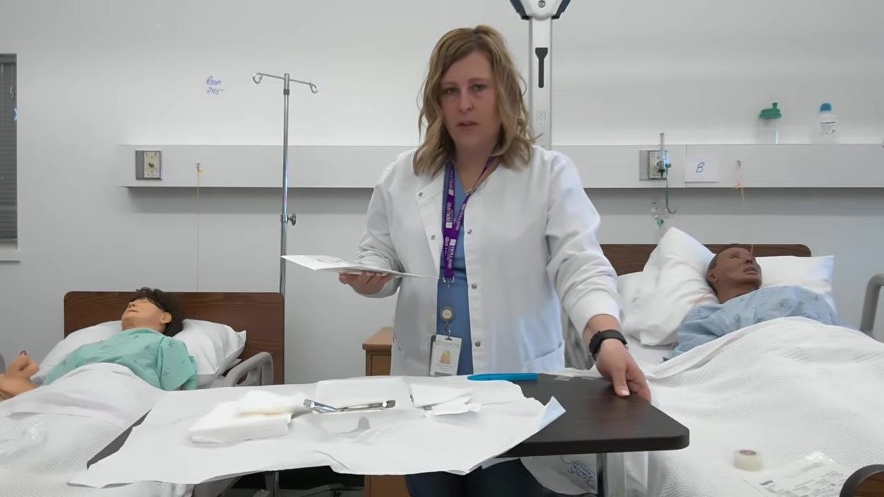 A healthcare professional moves the tray to a more convenient position while holding the glove package in the other hand. The sterile field remains organized, with medical mannequins visible in the background.