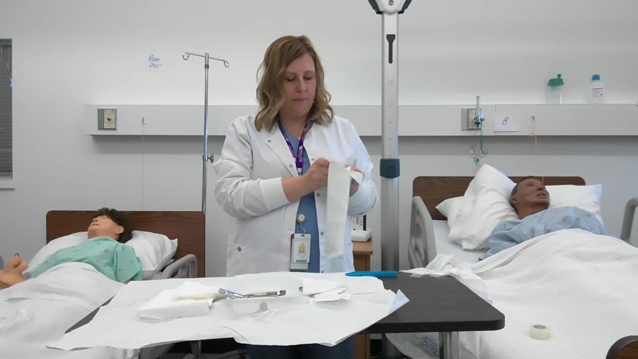 A healthcare professional opens the sterile glove package on the tray, preparing for gloving. The sterile field is visible with forceps, gauze, and other supplies.