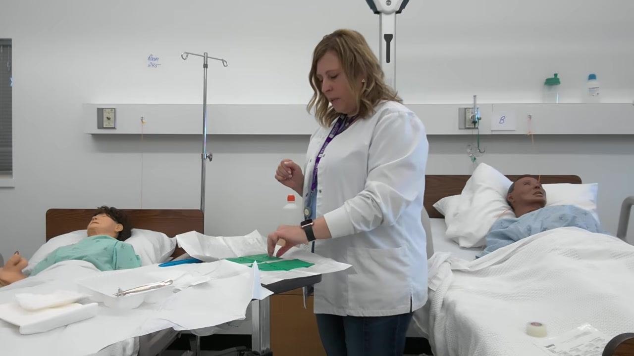 A healthcare professional prepares to pick up the first glove by the cuff, with green gloves visible on the sterile field.