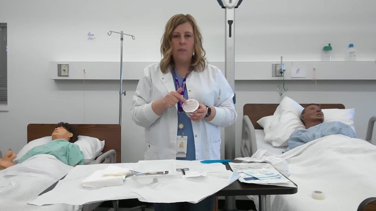A healthcare professional holds a container of saline solution and points to the label to check the expiration date, with the sterile tray and supplies visible.