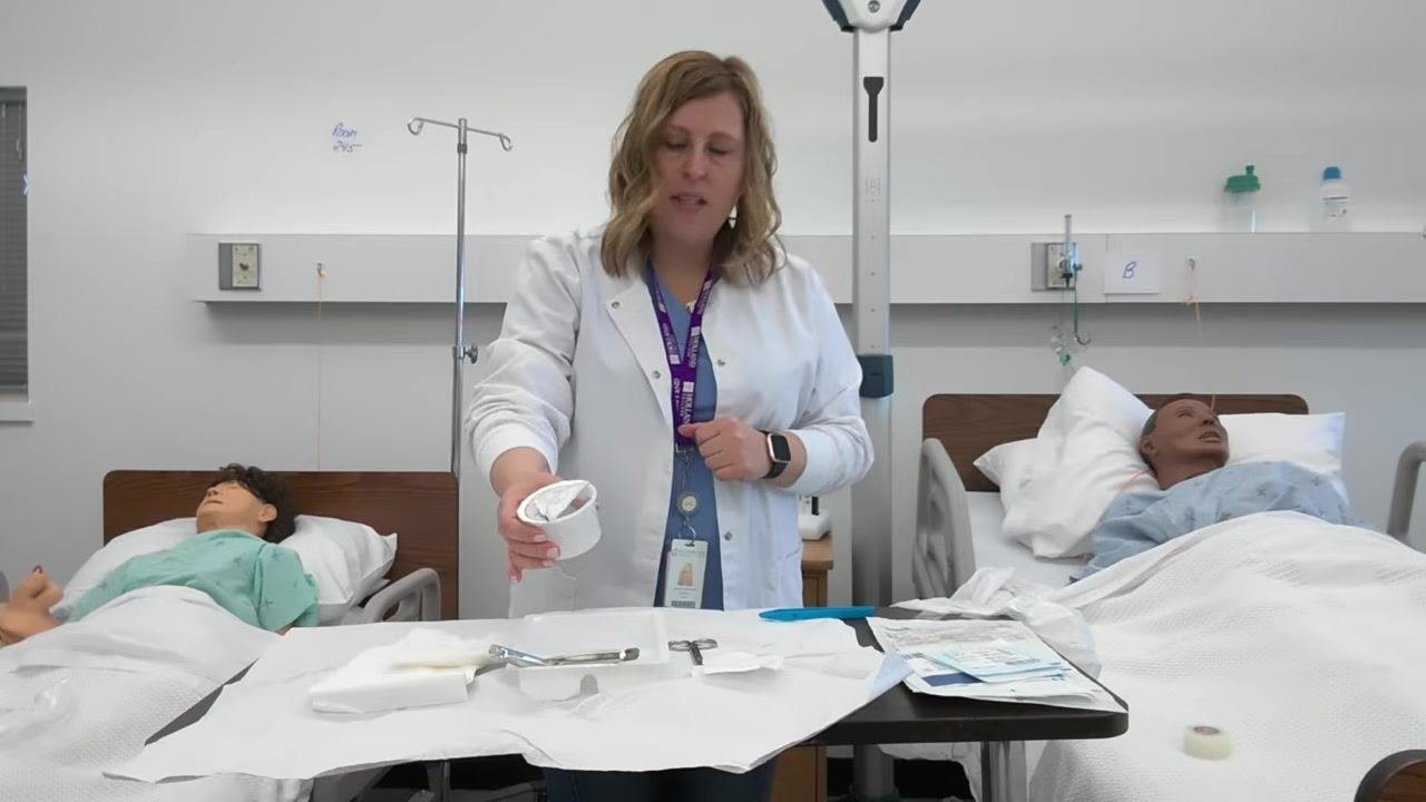 A healthcare professional holds an empty saline container above a sterile tray containing forceps, scissors, gauze, and tape, preparing to discard it into the garbage bag taped to the table.