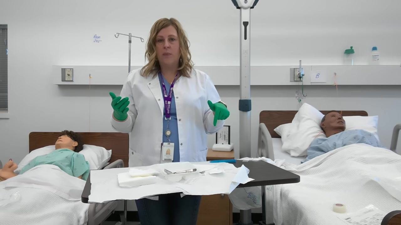 A healthcare professional with gloved hands raised stands between two hospital beds with mannequins. The sterile tray in front of her contains gauze, forceps, and other supplies, with used wrappers visible and being moved away.