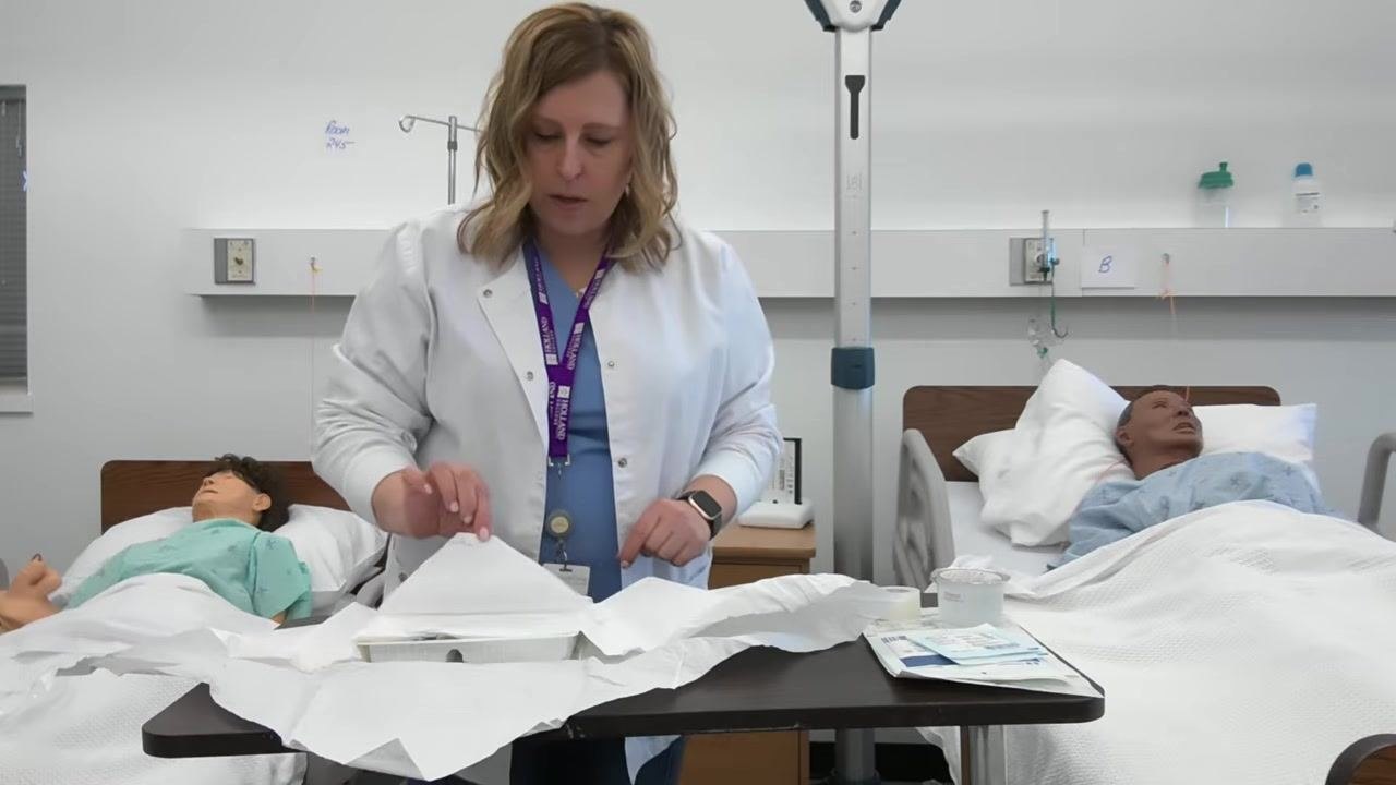 A healthcare professional reaches into the opened dressing tray to retrieve a set of blue forceps, in a clinical skills lab with two patient mannequins in hospital beds.