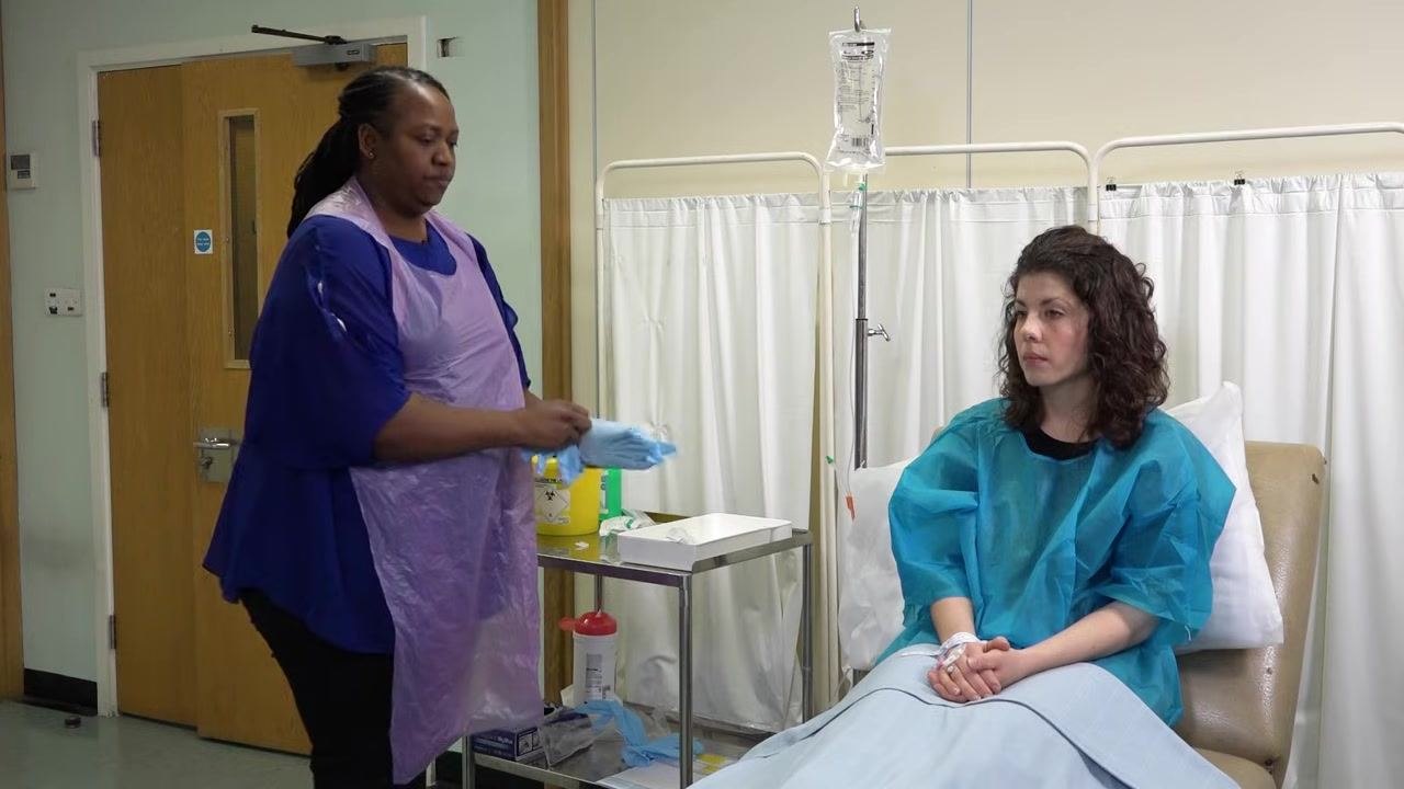 A staff member in a purple apron puts on a new pair of gloves, standing next to a tray with supplies and the patient.