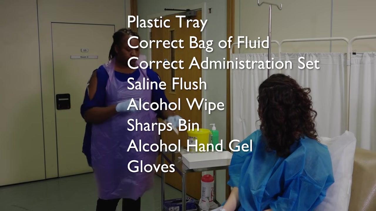 A staff member in a purple apron and gloves stands by a trolley with supplies. The equipment list is overlaid: Plastic Tray, Correct Bag of Fluid, Correct Administration Set, Saline Flush, Alcohol Wipe, Sharps Bin, Alcohol Hand Gel, Gloves.
