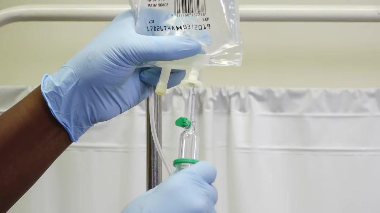 A staff member in gloves and a purple apron carefully unravels the IV administration set, holding both ends to maintain sterility.