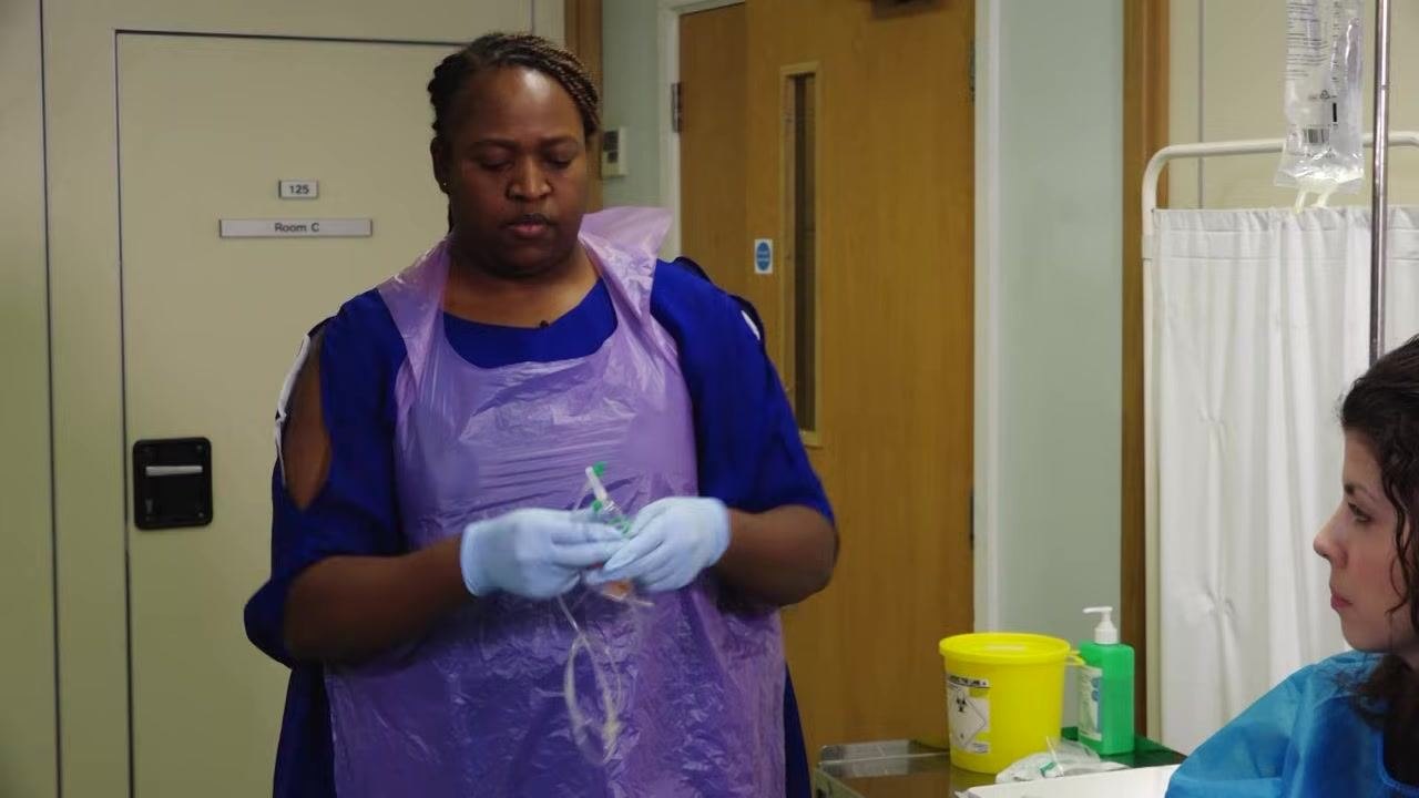 A staff member in gloves and a purple apron removes the cap from the spike of the IV administration set, holding it carefully to maintain sterility.