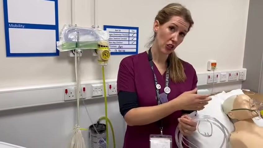 A healthcare professional in scrubs stands beside a hospital bed, holding a nasal cannula and explaining the procedure. The patient mannequin is on the bed, and oxygen equipment is visible on the wall.