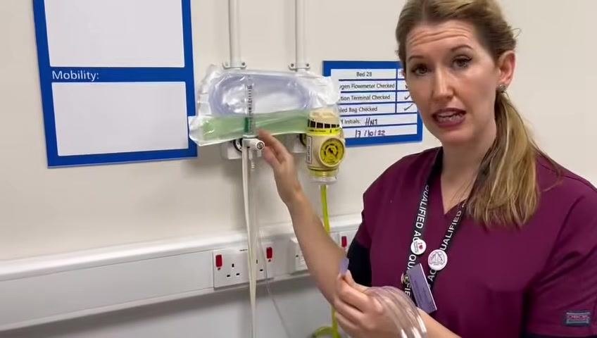 The healthcare professional points to and adjusts the oxygen flow meter on the wall, with a Venturi mask and tubing visible above.