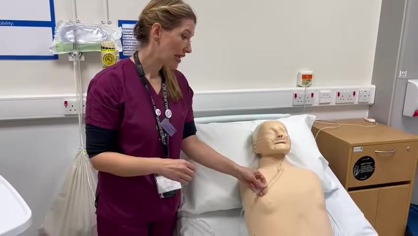 The healthcare professional stands beside the mannequin patient, holding the nasal cannula tubing and discussing flow rate adjustments.