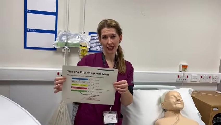 The healthcare professional concludes the demonstration, holding the oxygen conversion chart and standing beside the mannequin patient.
