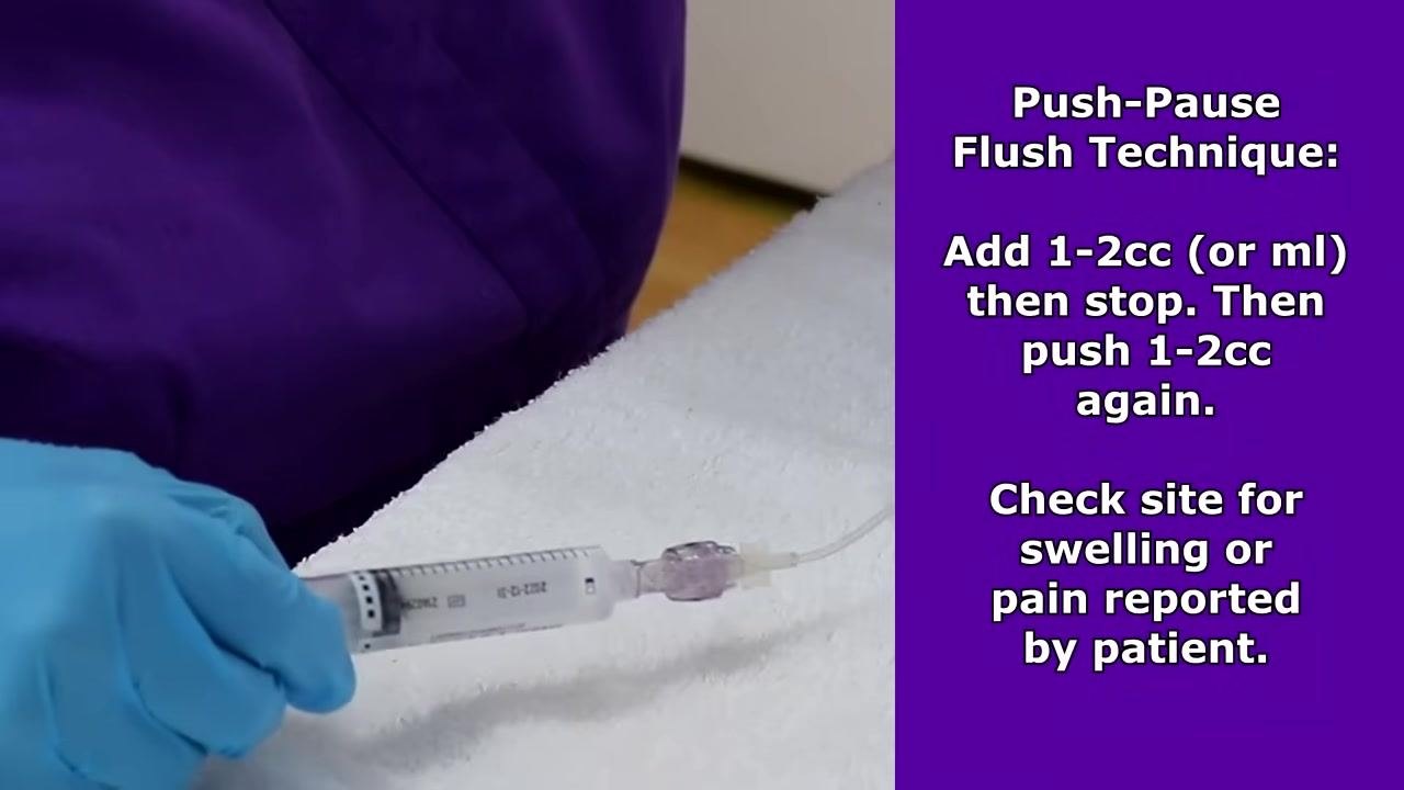 Holding a syringe attached to an IV extension set while demonstrating the push-pause flush technique, with on-screen text explaining the procedure and instructing to check the site for swelling or reported pain