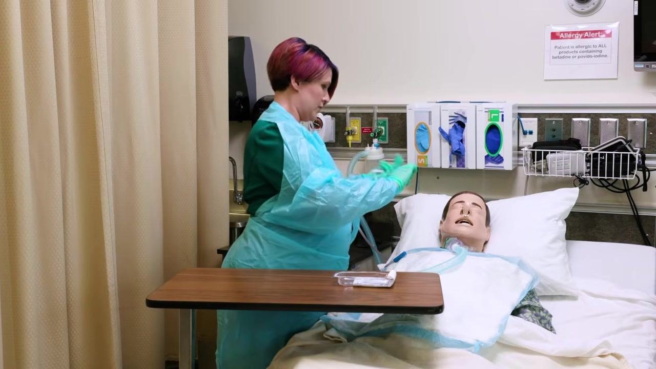 Clinician in PPE approaching mannequin patient, holding suction catheter above patient, maintaining sterile technique