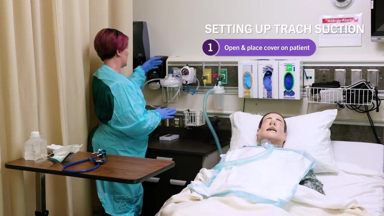 Close-up of nurse in blue gown and gloves holding suction tubing, pointing to the suction regulator gauge, with a label indicating "100-150 mm HG for adults"