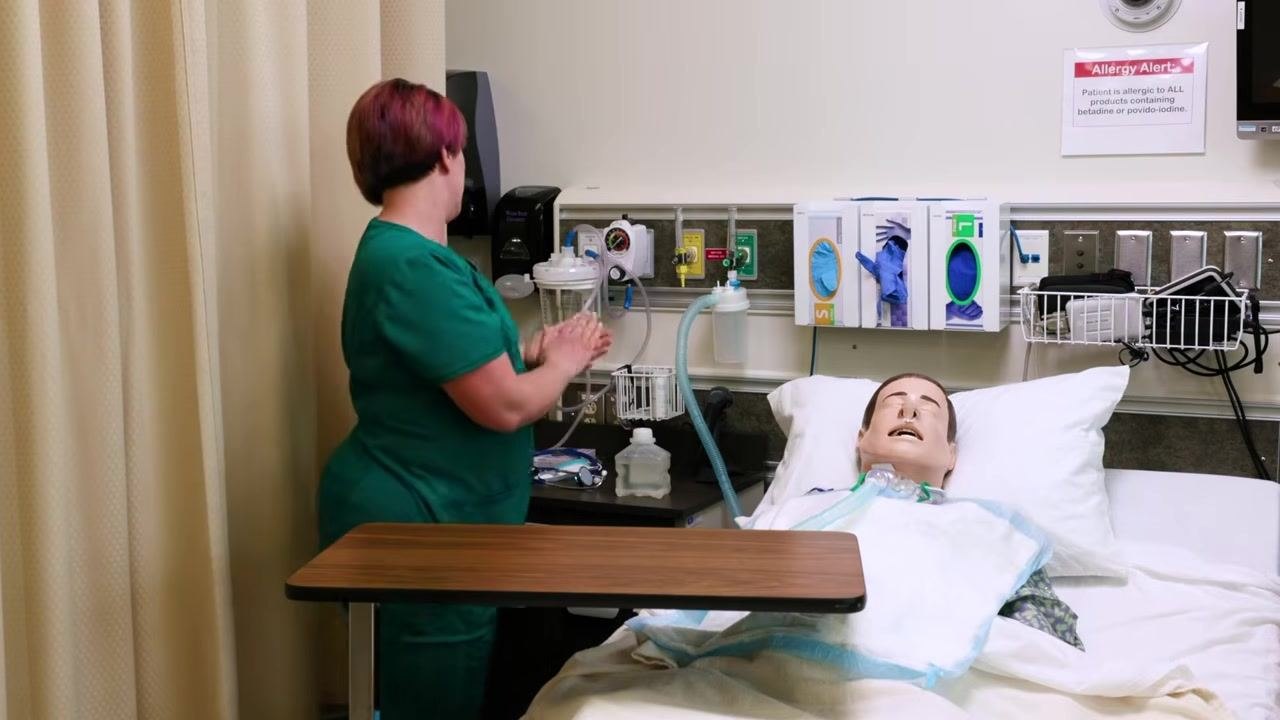 Clinician in scrubs, back to camera, performing hand hygiene at sink after removing PPE. Mannequin patient remains in bed