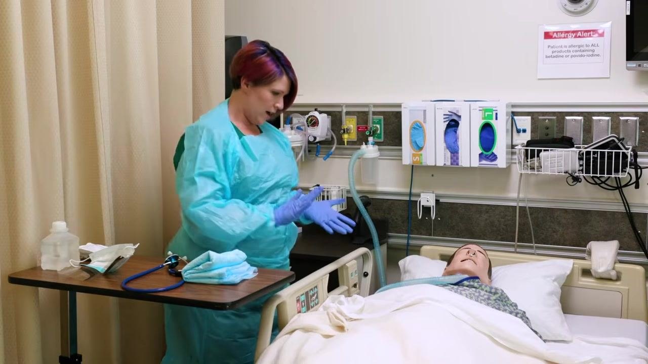 Nurse in blue gown and gloves adjusting the bed rail of a mannequin patient, with PPE and medical equipment visible on the wall