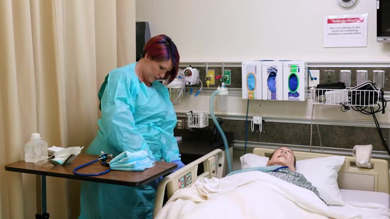 Nurse in blue gown and gloves using a stethoscope to assess the mannequin patient's lung sounds, with PPE and medical equipment visible on the wall