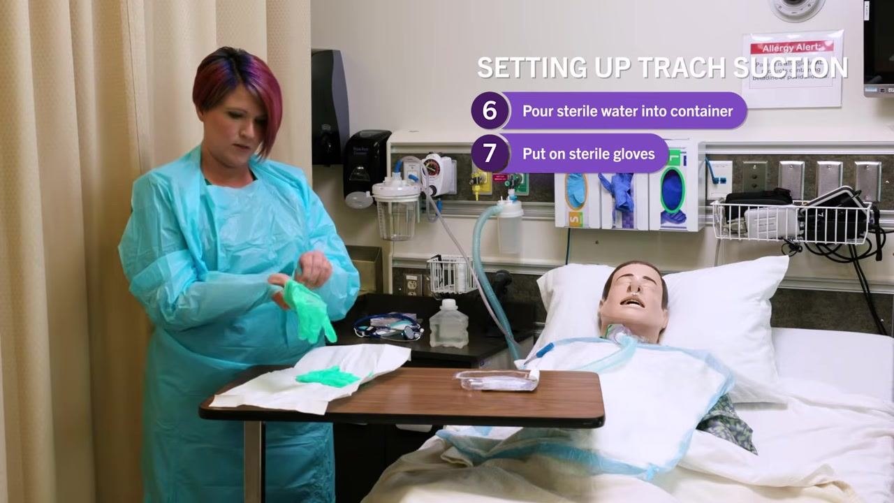 Nurse in blue gown putting on green sterile gloves at the bedside table, with the sterile field and supplies visible. On-screen checklist: "7. Put on sterile gloves"