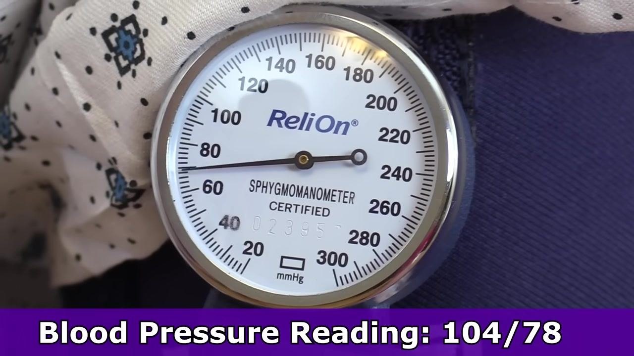 Close-up of a sphygmomanometer gauge showing the needle at 104 mmHg, labeled "ReliOn"