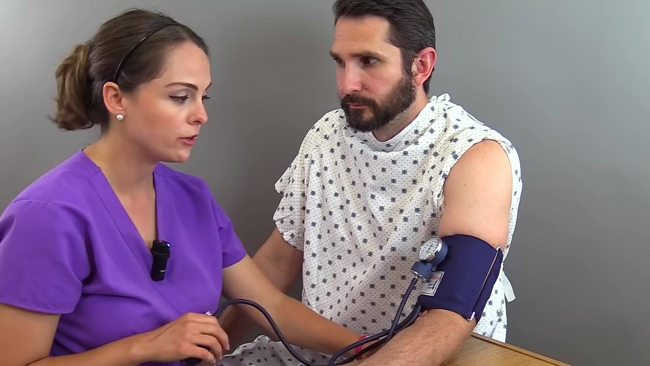 Nurse positioning the cuff about two inches above the bend of the patient's left arm, with the arrow on the cuff visible and the patient holding their sleeve up