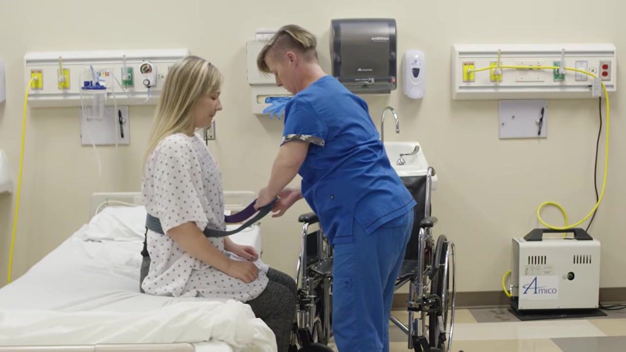 The healthcare worker is securing a transfer (gait) belt around the client's waist while the client sits on the edge of the bed, ready for transfer.