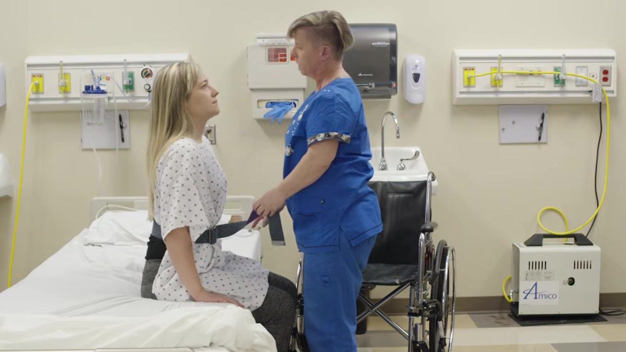 A healthcare worker in blue scrubs stands directly in front of a client sitting on the edge of a hospital bed, holding the transfer belt and preparing to assist the client to stand. The environment includes a wheelchair positioned next to the bed, a hospital sink, and medical equipment on the wall.