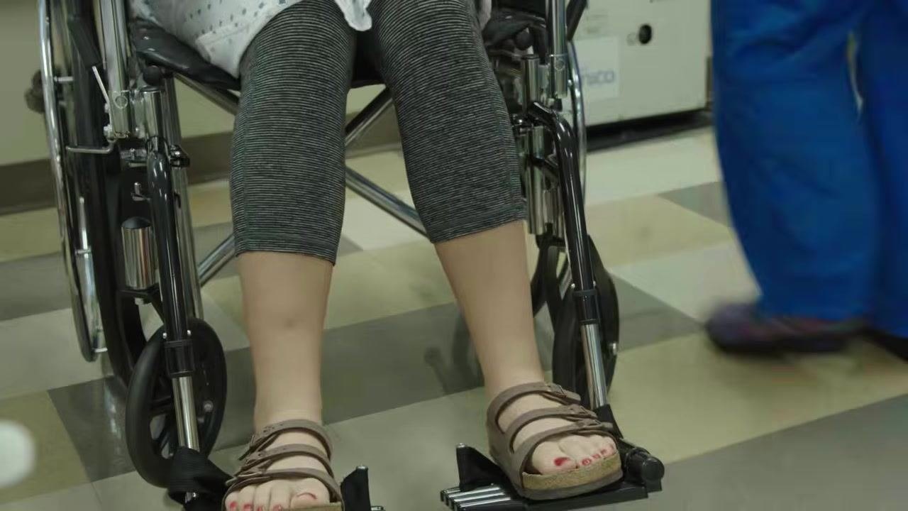 The healthcare worker is bent over, removing the transfer belt from the client who is now seated in the wheelchair. The client is positioned with her hips against the back of the wheelchair.