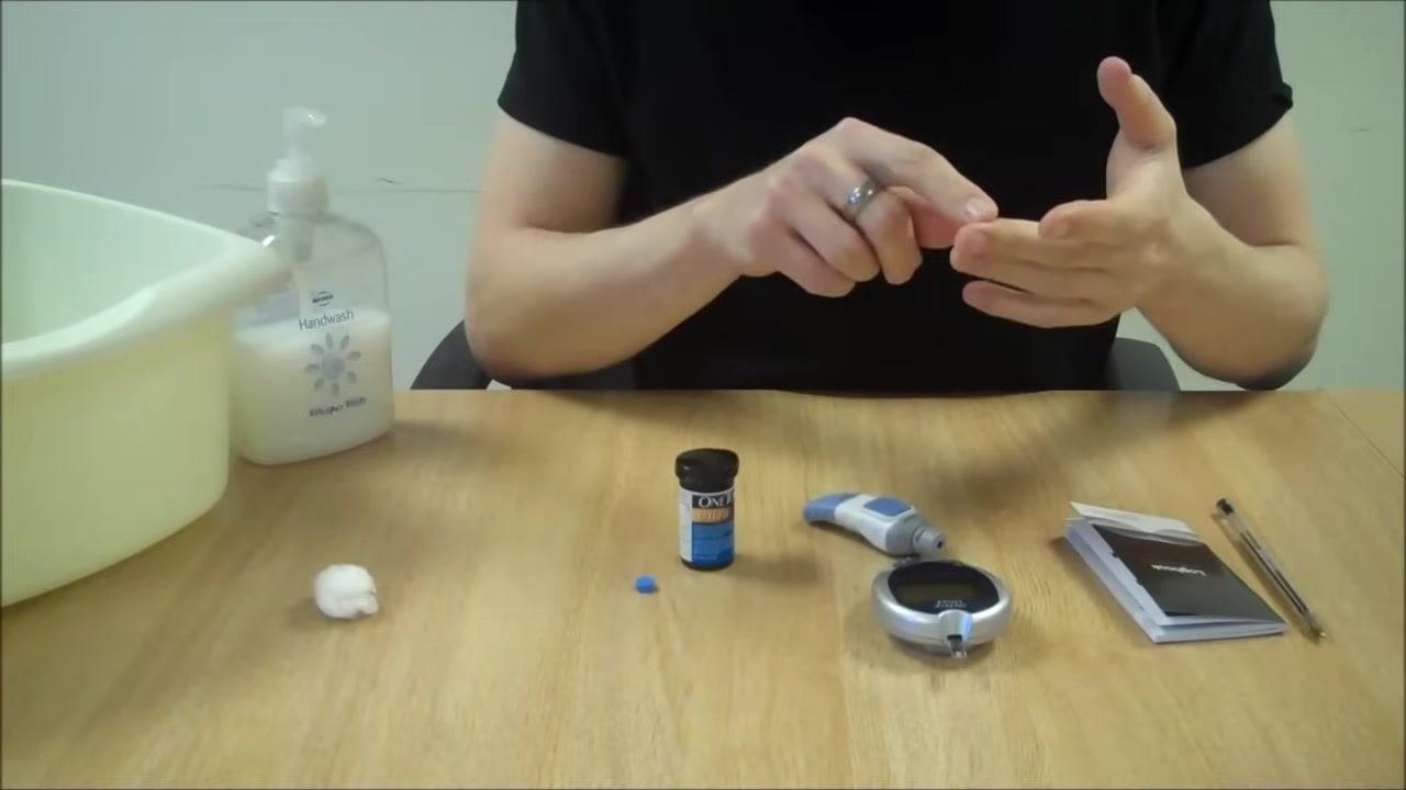 Person indicating the side of their finger, preparing for the finger prick, with the meter and lancing device on the table