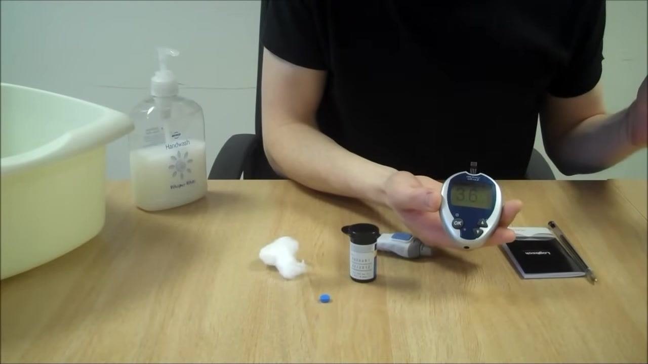 Person holding the blood glucose meter with the result displayed as 3.6 mmol/L, with all testing equipment visible on the table: handwash, bowl, cotton wool, test strip container, lancing device, monitoring diary, and pen
