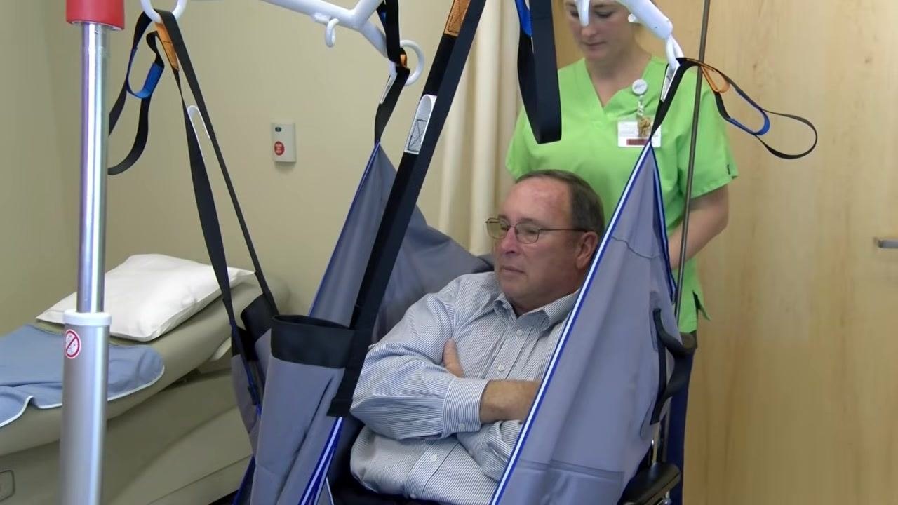 The patient is lowered onto the exam table while one healthcare worker guides the lift and another adjusts the sling for a safe landing.