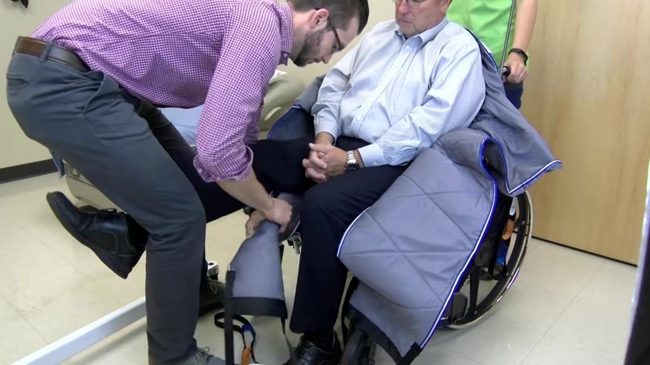 A caregiver lifts the patient's leg and pulls the sling underneath it while the patient sits in the wheelchair.